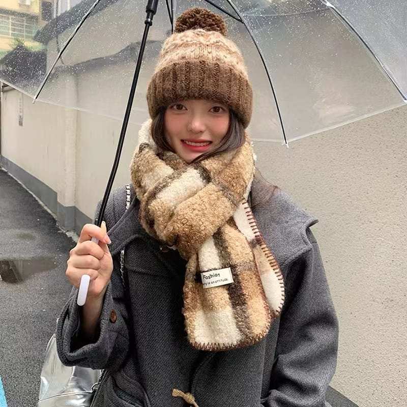 Woman wearing brown cream plaid sherpa scarf and chunky knit pom-pom beanie hat holding clear umbrella in rain