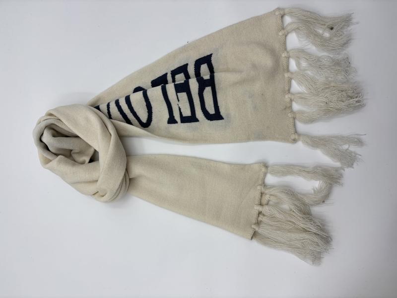 Cream knit scarf with navy blue varsity-style letter print and tassel fringe ends, laid flat on white background