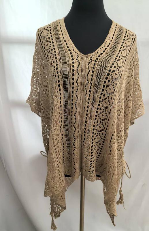 Beige open-weave crochet poncho top with floral lace pattern, V-neckline, short sleeves, and tassel fringe hem on mannequin