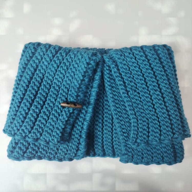 Folded teal chunky knit cowl neck warmer with wooden toggle button closure, ribbed texture, displayed on white surface