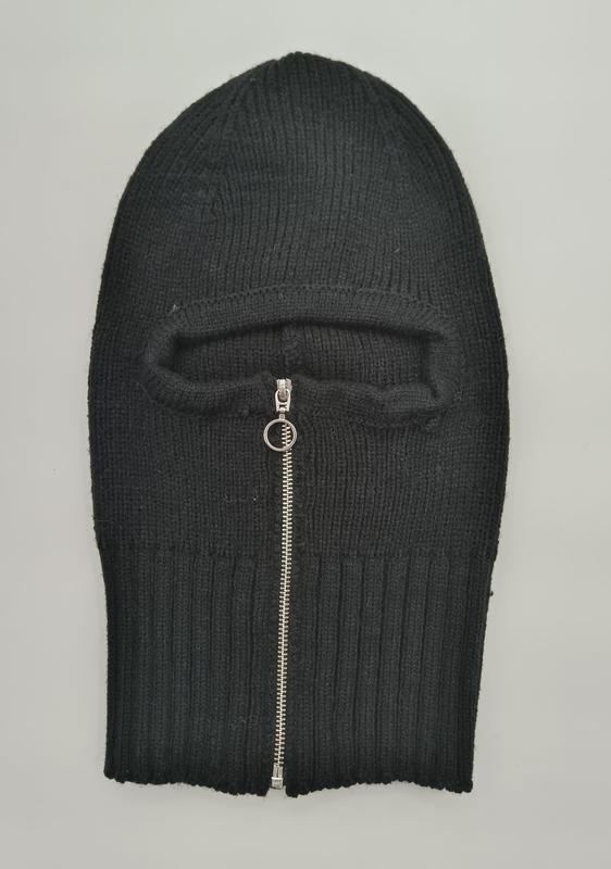 Black ribbed knit balaclava laid flat showing a silver zipper opening for the face area on a grey background