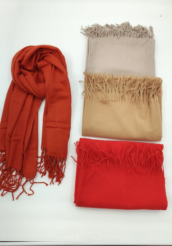 Four pashmina scarves with fringe in rust orange, light beige, camel, and red, one draped and three folded on white surface