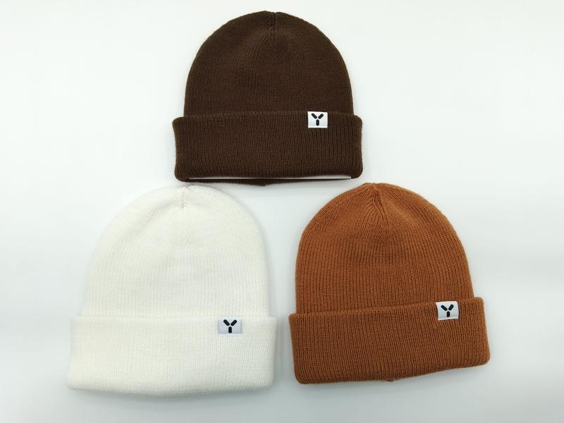Three cuffed knit beanies laid flat in dark brown, cream white, and caramel orange, each with a small logo patch label