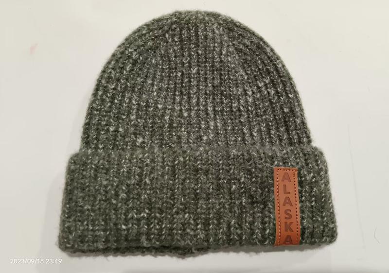 Olive green chunky ribbed knit beanie hat with folded cuff and brown leather Alaska patch label on white background
