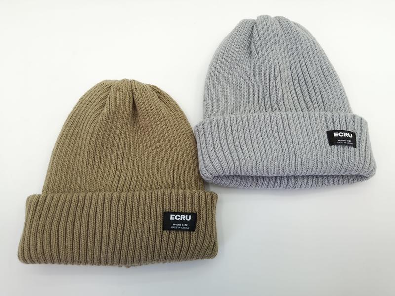 Two ribbed knit cuffed beanies side by side in khaki and light grey with black woven brand label on folded cuff