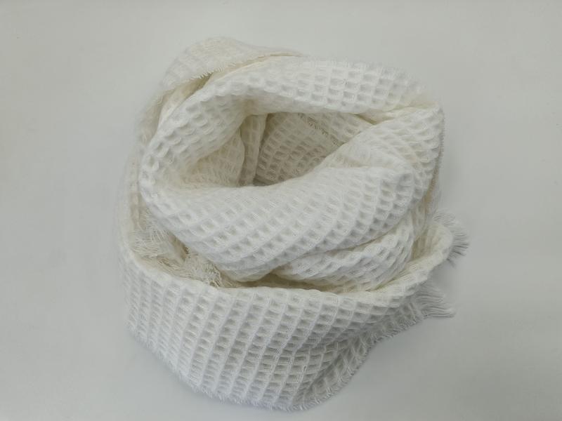 White waffle texture knit scarf coiled on white surface showing fringe edges and open weave pattern