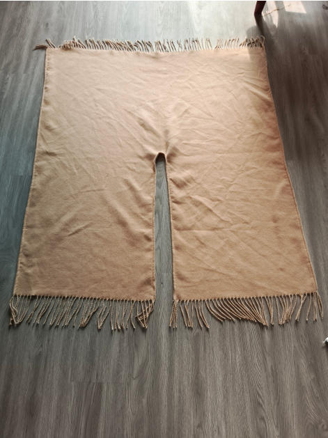 Flat lay of a beige woven poncho with center neck slit and twisted fringe along top and bottom edges on wood floor