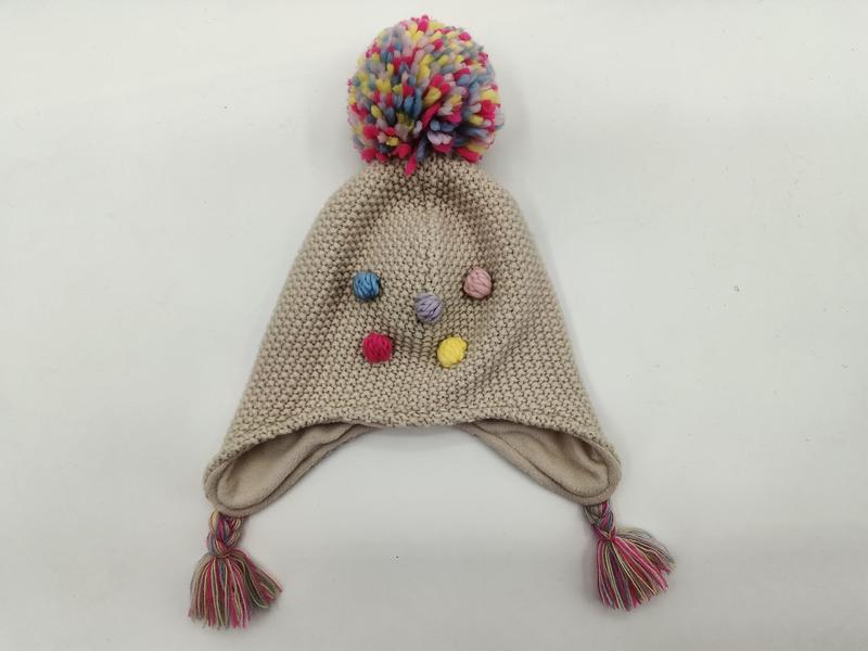 Beige knitted children's earflap hat with large multicolor pom-pom on top, colorful bobble accents, and braided tassels
