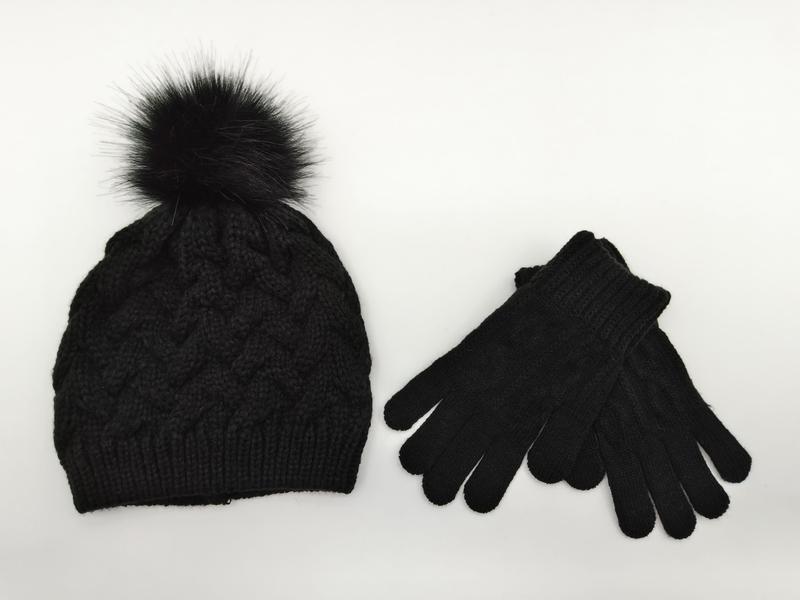Black cable knit winter beanie hat with faux fur pom-pom and matching knit gloves set on white background