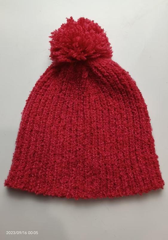 Red chunky ribbed knit winter beanie hat with large fluffy pom-pom on top, laid flat on white background