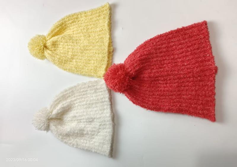 Three knitted pom-pom beanies in yellow, red, and white laid flat on white surface, fuzzy winter hats