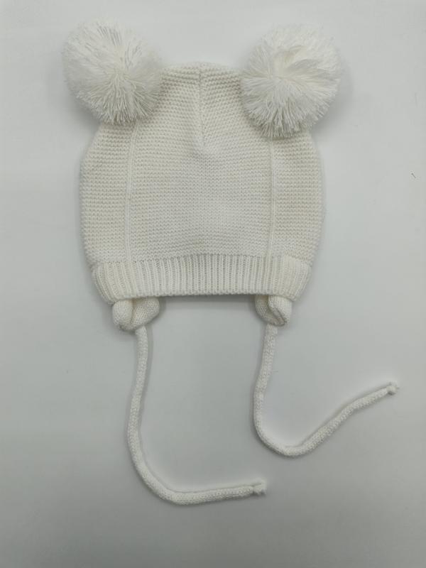 White knitted baby beanie with two fluffy pom poms on top and long chin tie strings, laid flat on white background
