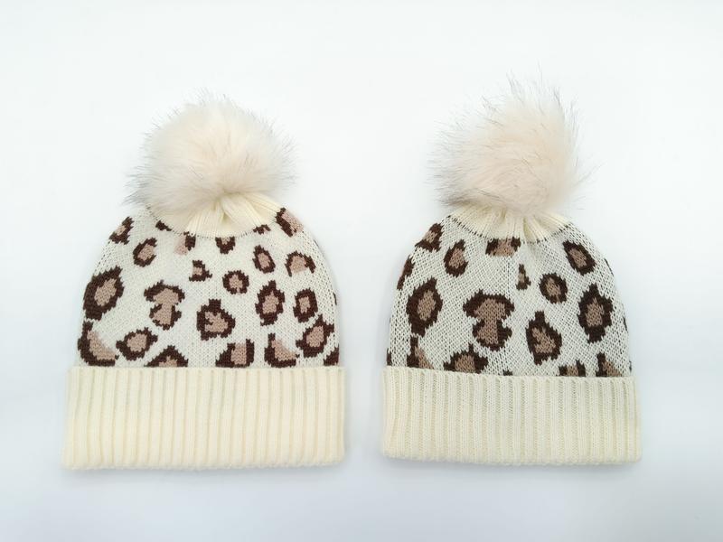 Two cream ivory leopard print knit beanies with fluffy faux fur pom-poms, adult and child sizes on white background