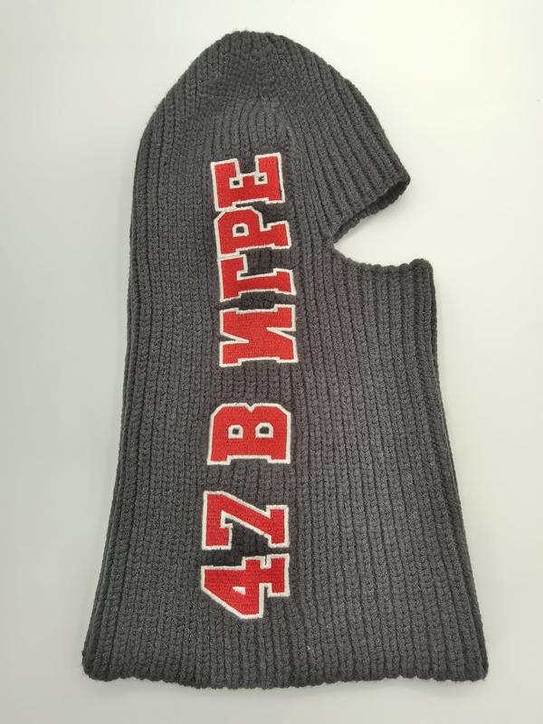 Gray ribbed knit balaclava ski mask laid flat with bold red and white embroidered text on dark charcoal wool fabric