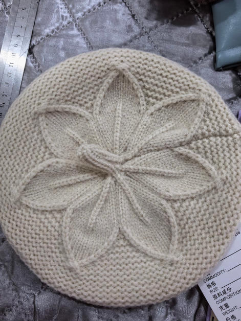 Cream knitted beret hat with raised floral leaf cable pattern at crown, laid flat on silver fabric