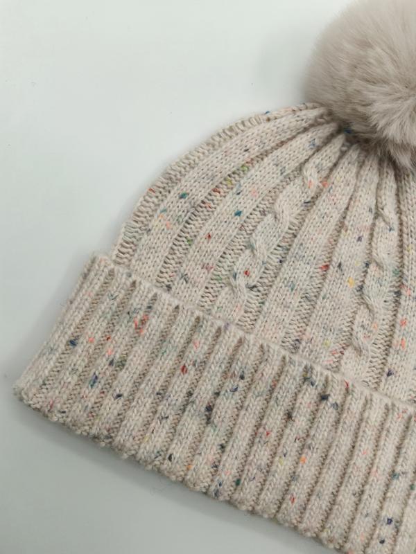 Close-up of a cream cable knit beanie hat with multicolor fleck detailing, ribbed cuff, and fluffy faux fur pom pom on top