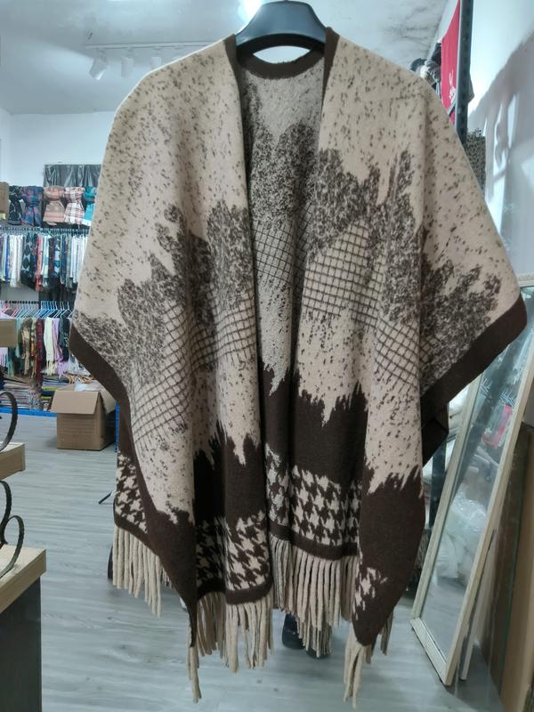 Beige and brown jacquard knit open-front poncho cape with floral pattern, houndstooth border, and fringe hem on hanger