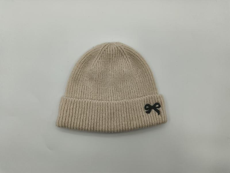 Cream ribbed knit beanie hat laid flat on white background featuring a small black embroidered bow on the folded cuff