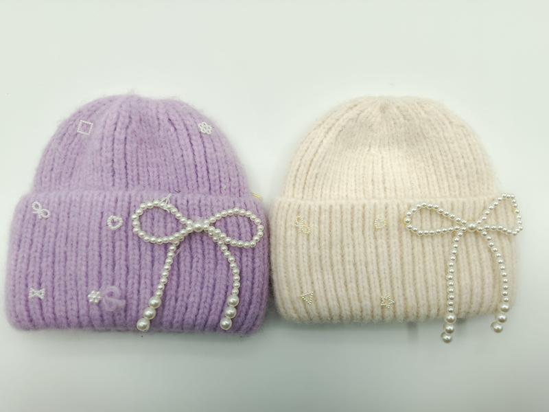 Two chunky knit beanies in lavender and cream with pearl bow decorations and embroidered motifs on white background