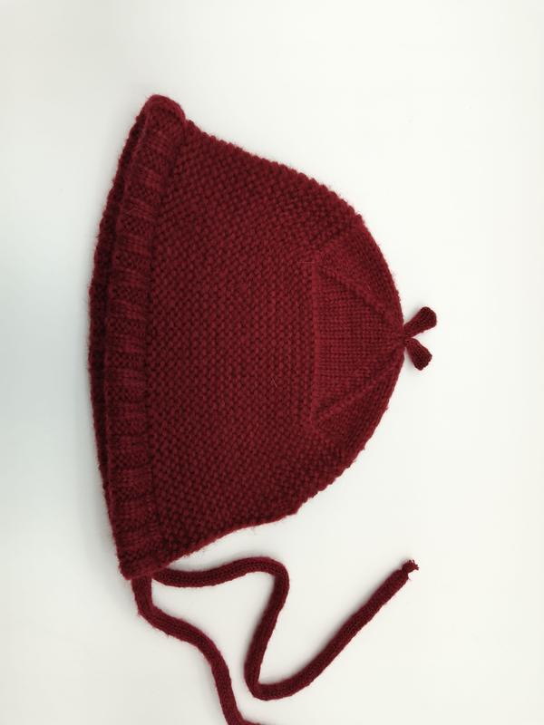 Hand-knitted dark red baby bonnet with garter stitch texture, ruffle edge, chin tie strings, and pointed top, laid flat on white background
